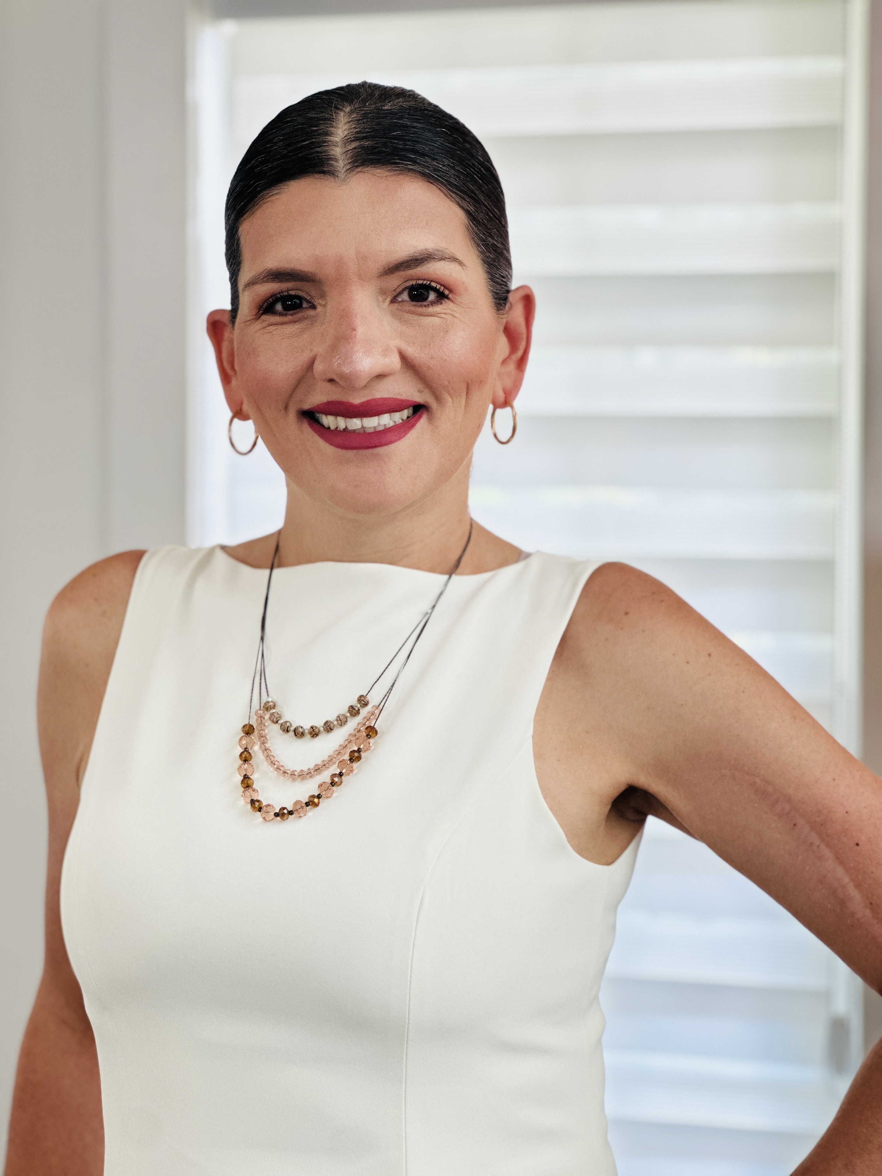 A woman with shoulder-length brown hair and a neutral expression rests her chin on her hand, wearing a light-colored blazer, with an out-of-focus seascape in the background.